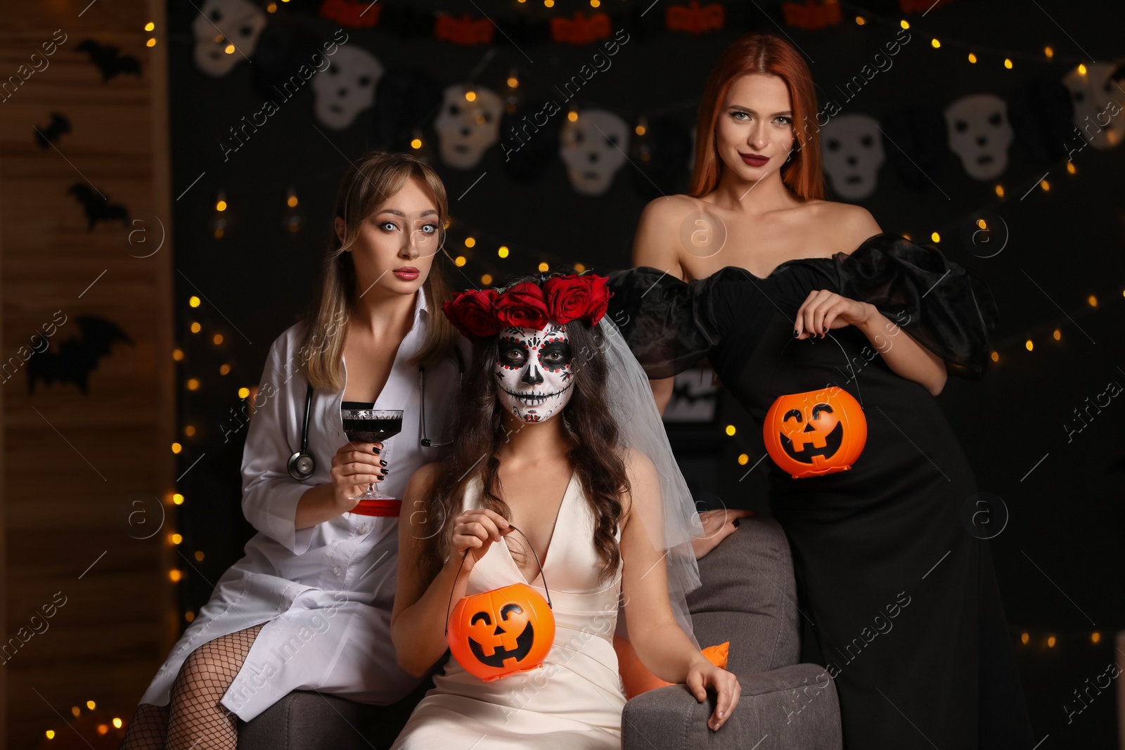 Photo of Group of women in scary costumes with pumpkin buckets and wine against blurred lights indoors. Halloween celebration