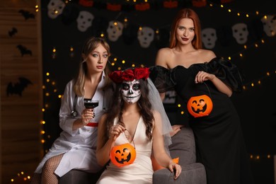 Group of women in scary costumes with pumpkin buckets and wine against blurred lights indoors. Halloween celebration