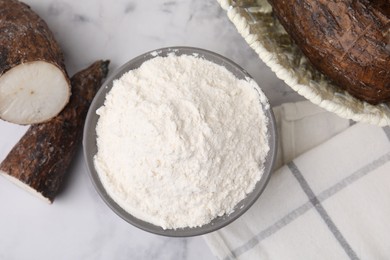 Photo of Bowl with cassava flour and roots on white marble table, flat lay