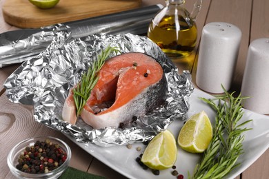 Photo of Aluminum foil with raw salmon, lime slices, rosemary and spices on wooden table, closeup