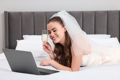 Photo of Happy bride with glass of champagne and laptop on bed in bedroom