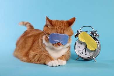 Cute ginger cat with sleep masks and alarm clock on light blue background