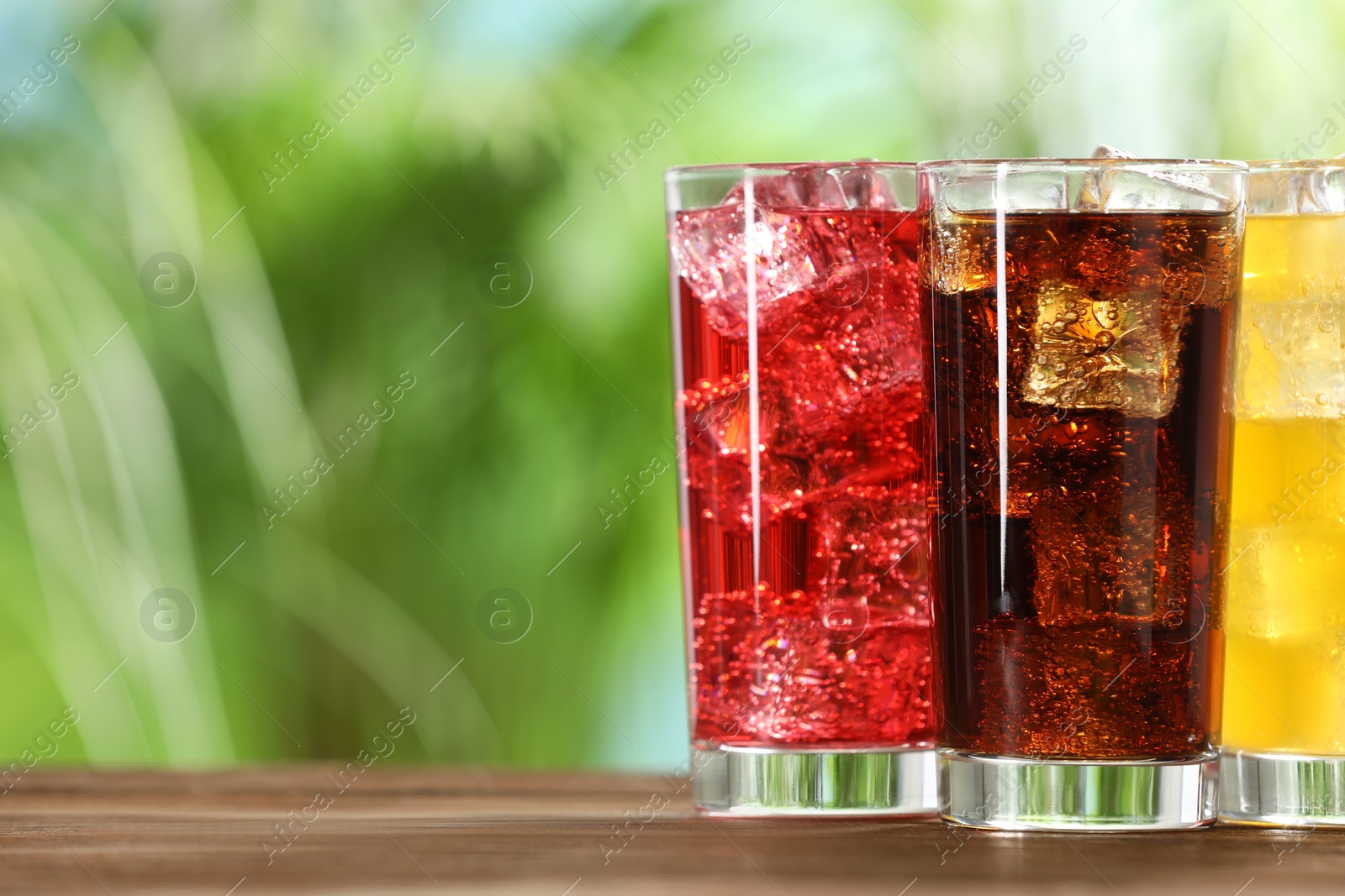 Photo of Glasses of different refreshing soda water with ice cubes on wooden table outdoors, space for text