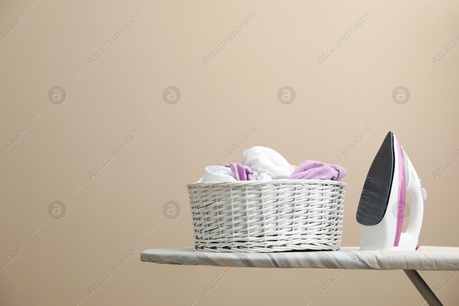 Photo of New modern iron and basket with laundry on board against beige background, space for text