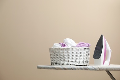 Photo of New modern iron and basket with laundry on board against beige background, space for text