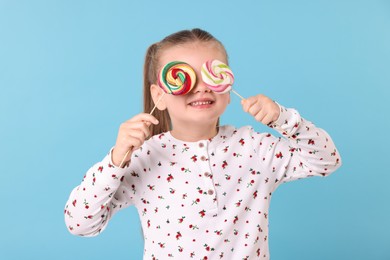 Happy little girl covering eyes with colorful lollipops on light blue background