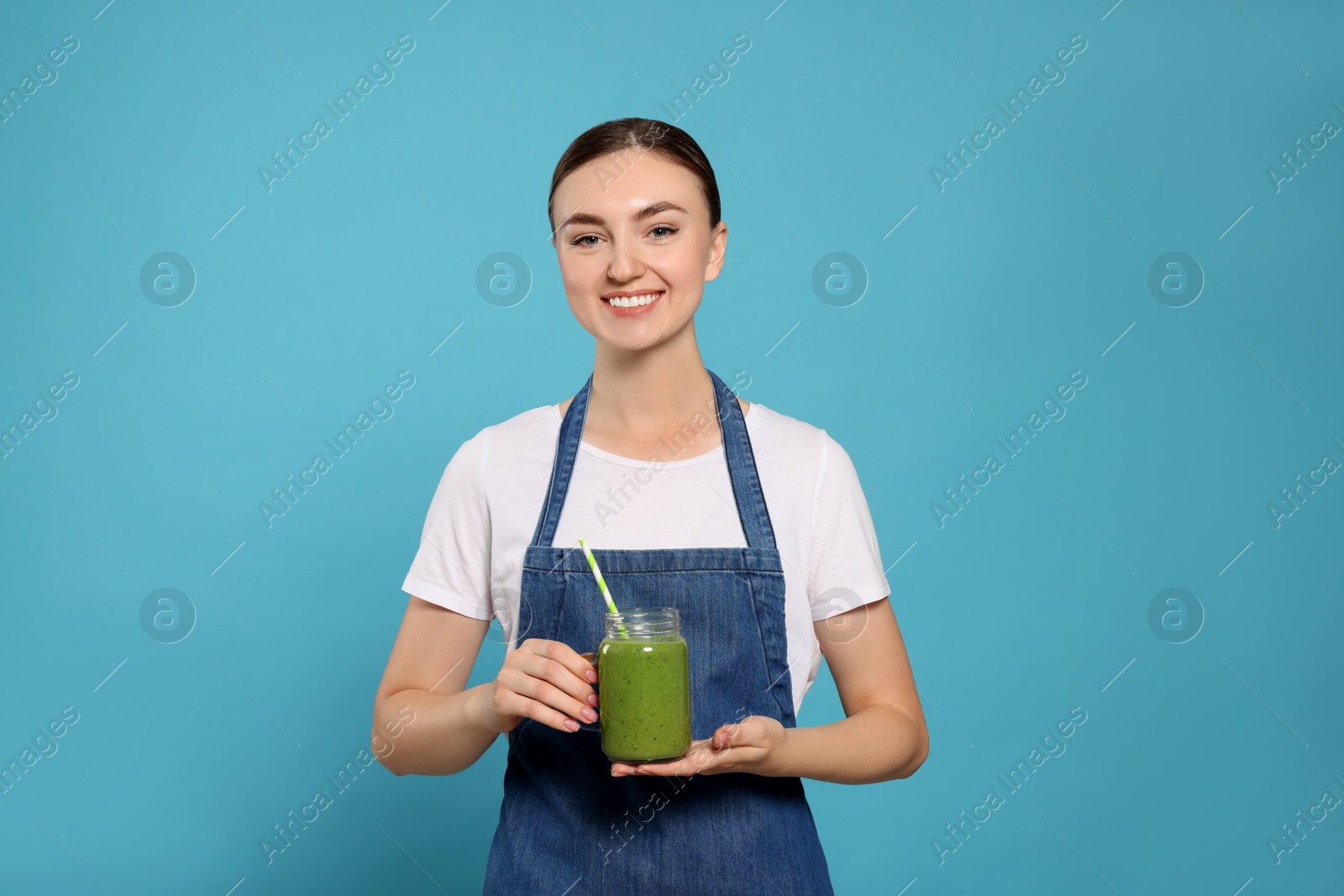 Photo of Beautiful young woman in denim apron with delicious smoothie on light blue background