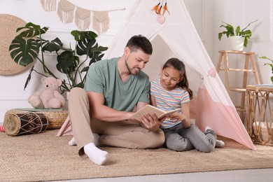Photo of Father and daughter reading book near toy wigwam at home