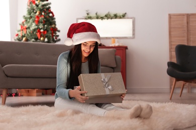 Photo of Beautiful young woman in Santa hat opening Christmas gift box at home