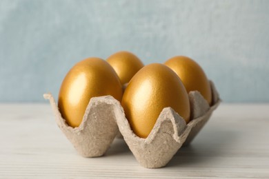 Carton with golden eggs on white wooden table, closeup