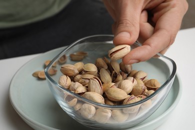 Woman taking tasty roasted pistachio nut from glass bowl at white table, closeup