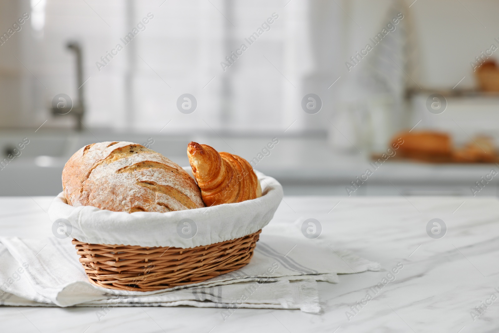 Photo of Wicker bread basket with freshly baked loaf and croissant on white marble table in kitchen, space for text