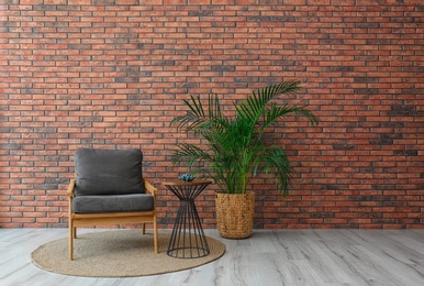 Modern room interior with stylish grey armchair and potted plant near brick wall