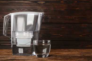 Filter jug and glass with purified water on wooden table. Space for text