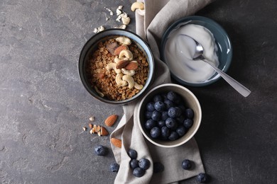 Photo of Tasty granola in bowl, blueberries, yogurt and spoon on gray textured table, flat lay. Space for text