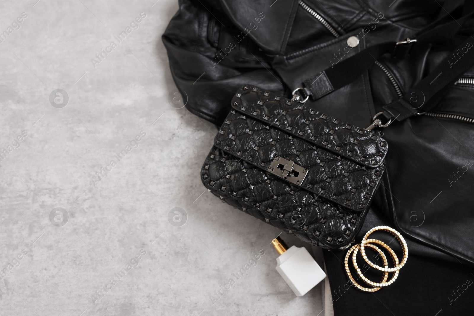 Photo of Bag, black leather jacket, bottle of perfume and golden bracelets on grey table, above view. Space for text