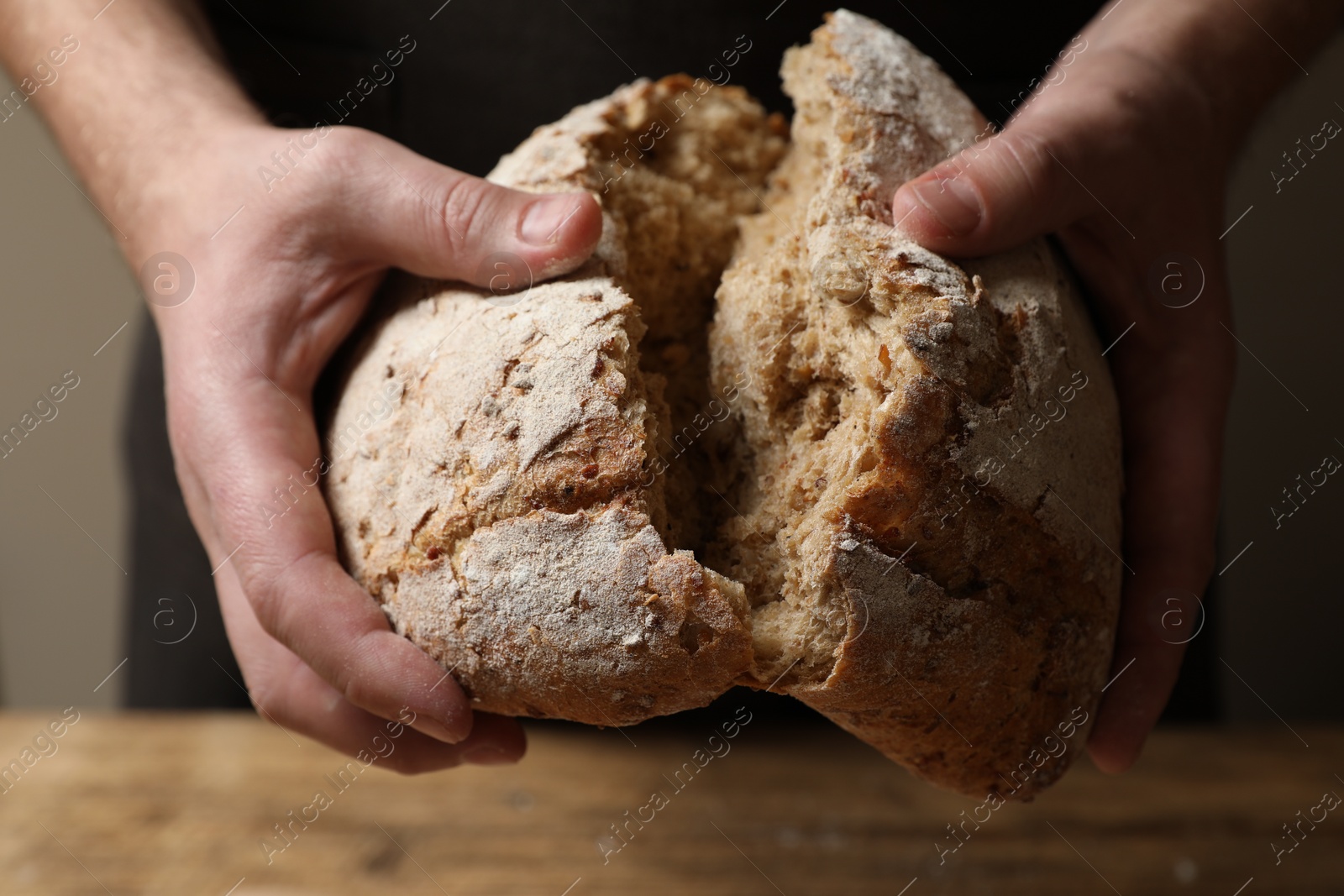 Photo of Man breaking loaf of fresh bread at wooden table, closeup