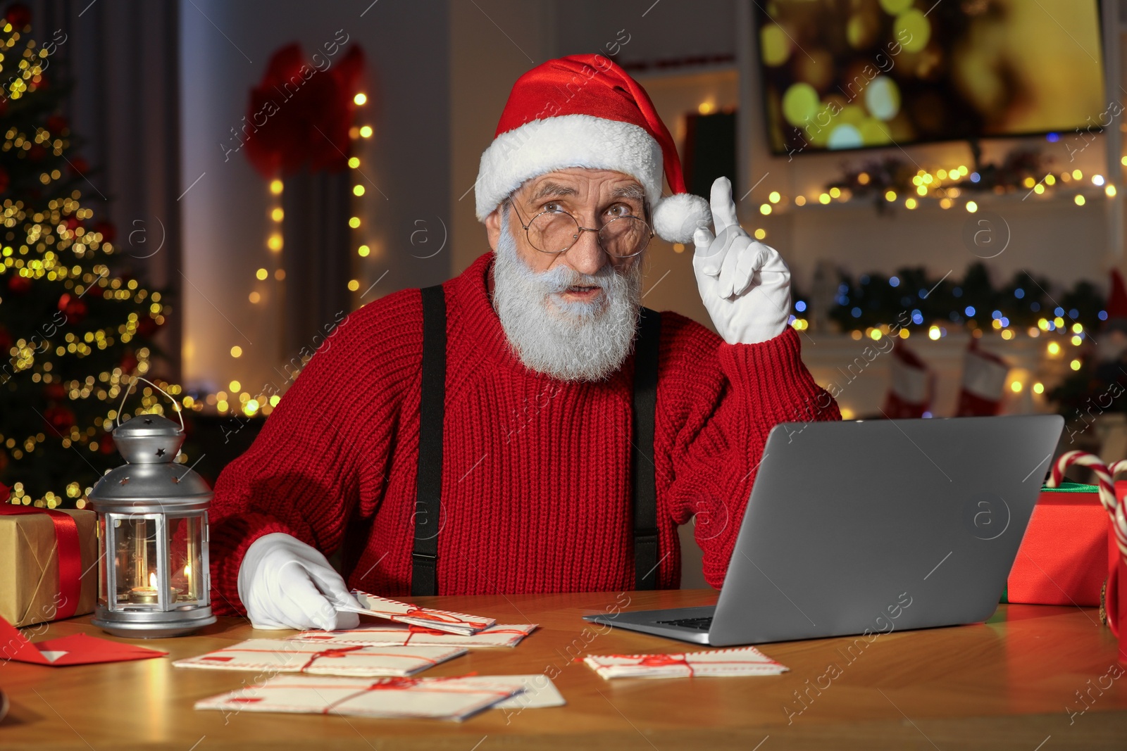 Photo of Santa Claus using laptop at his workplace in room decorated for Christmas