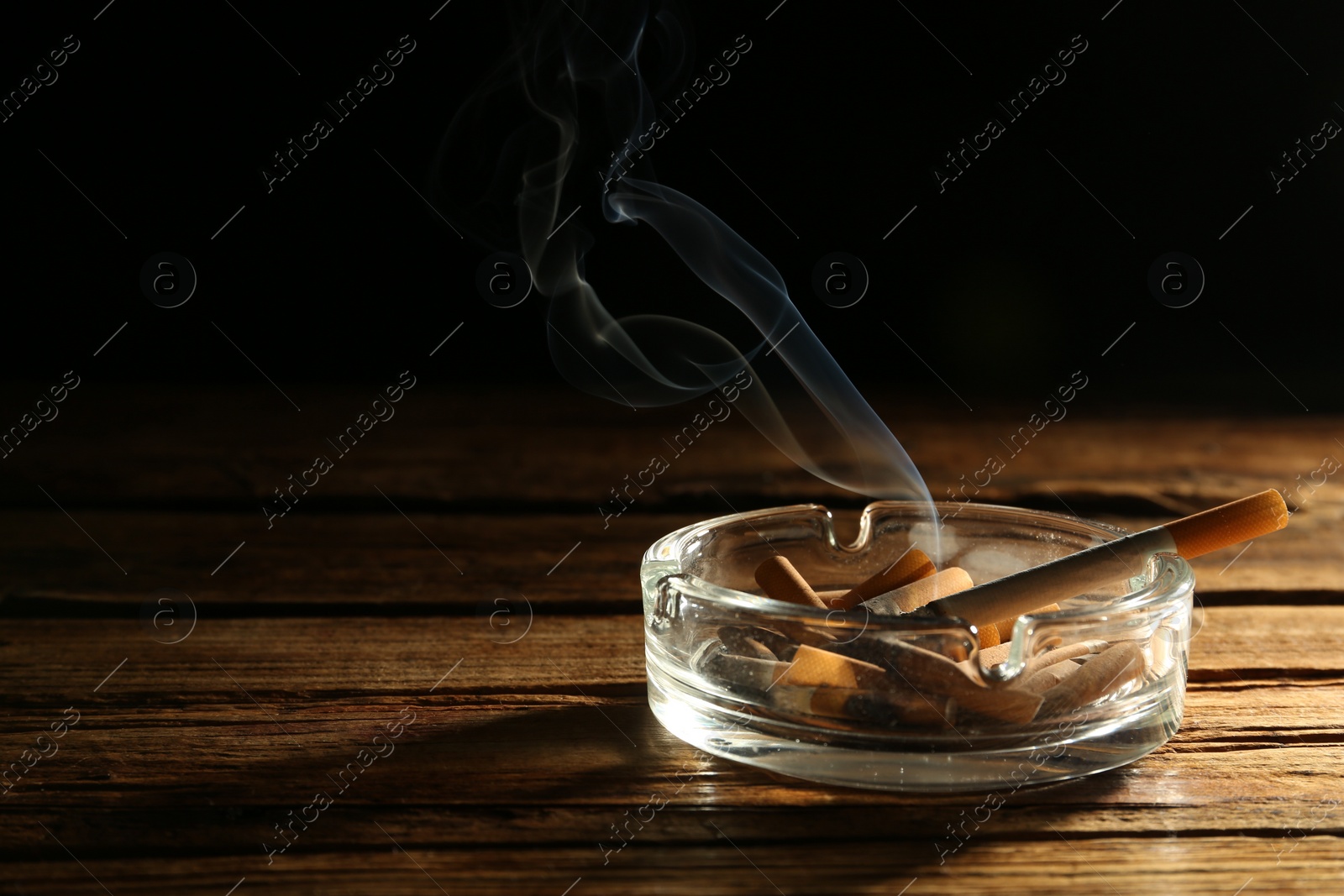 Photo of Smoldering cigarette in glass ashtray on wooden table against black background. Space for text