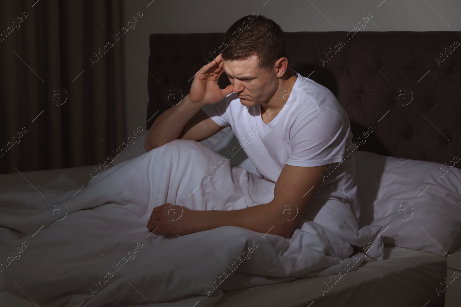 Photo of Handsome young man suffering from headache while sitting in bed at night