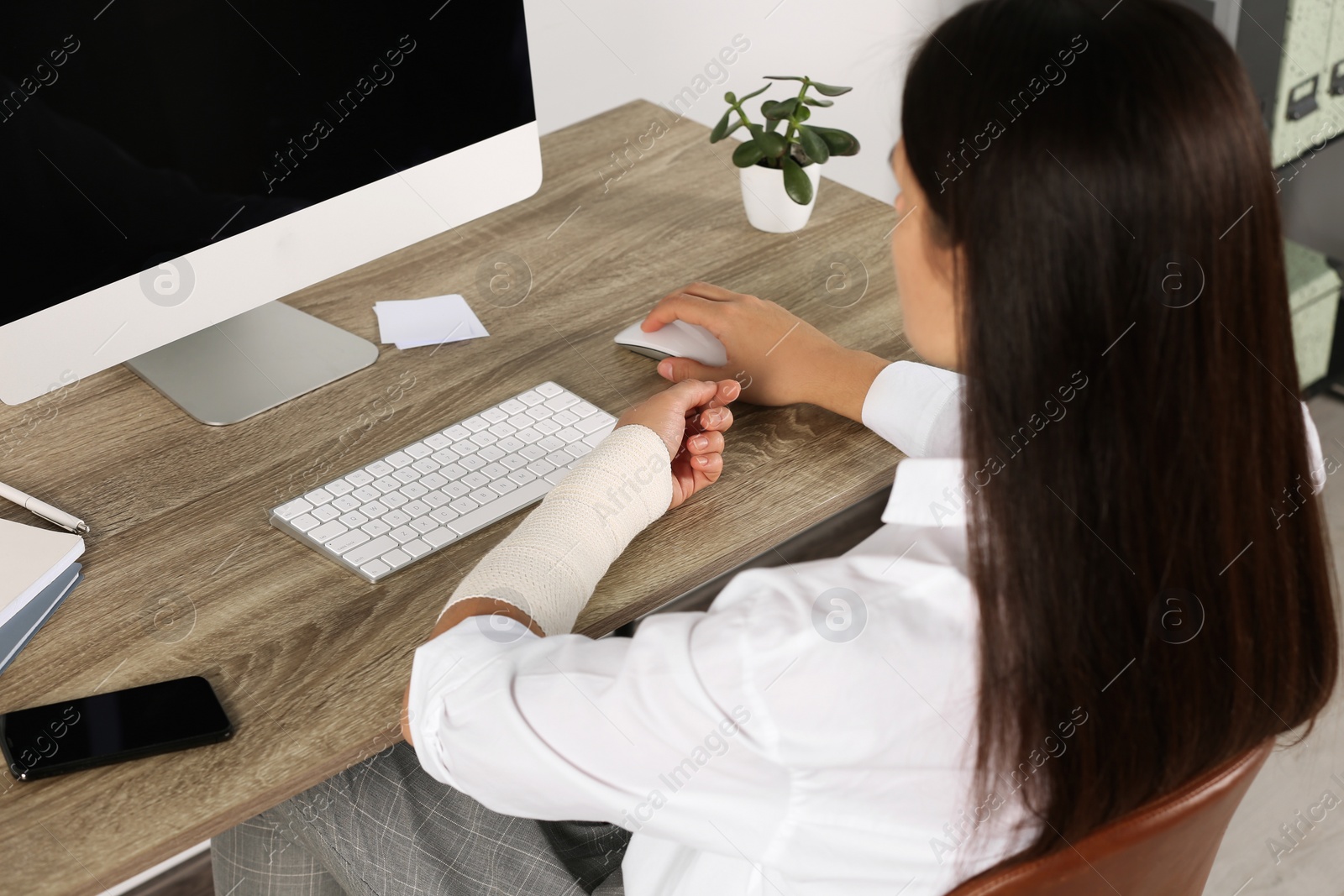 Photo of Young woman with arm wrapped in medical bandage sitting at workplace