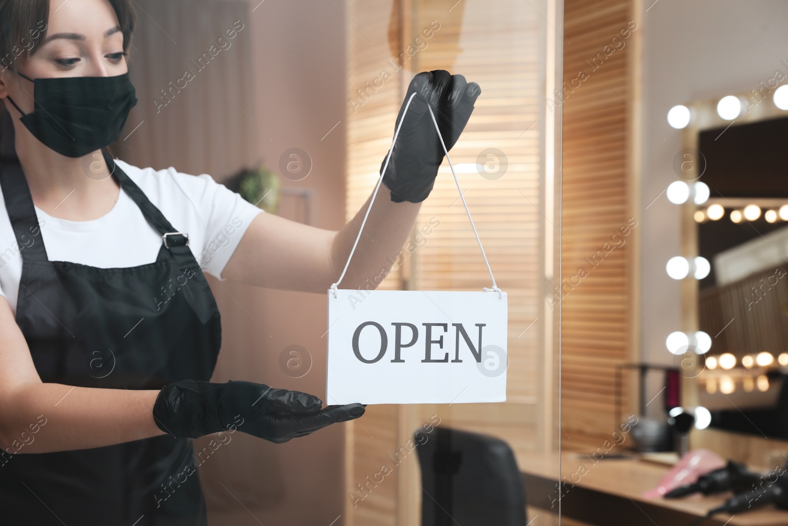 Photo of Woman with protective mask hanging Open sign onto glass door in salon. Beauty services during Coronavirus quarantine
