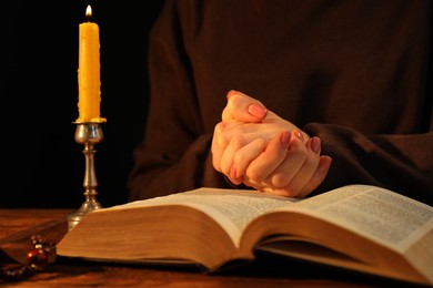 Woman praying at table with burning candle and Bible, closeup