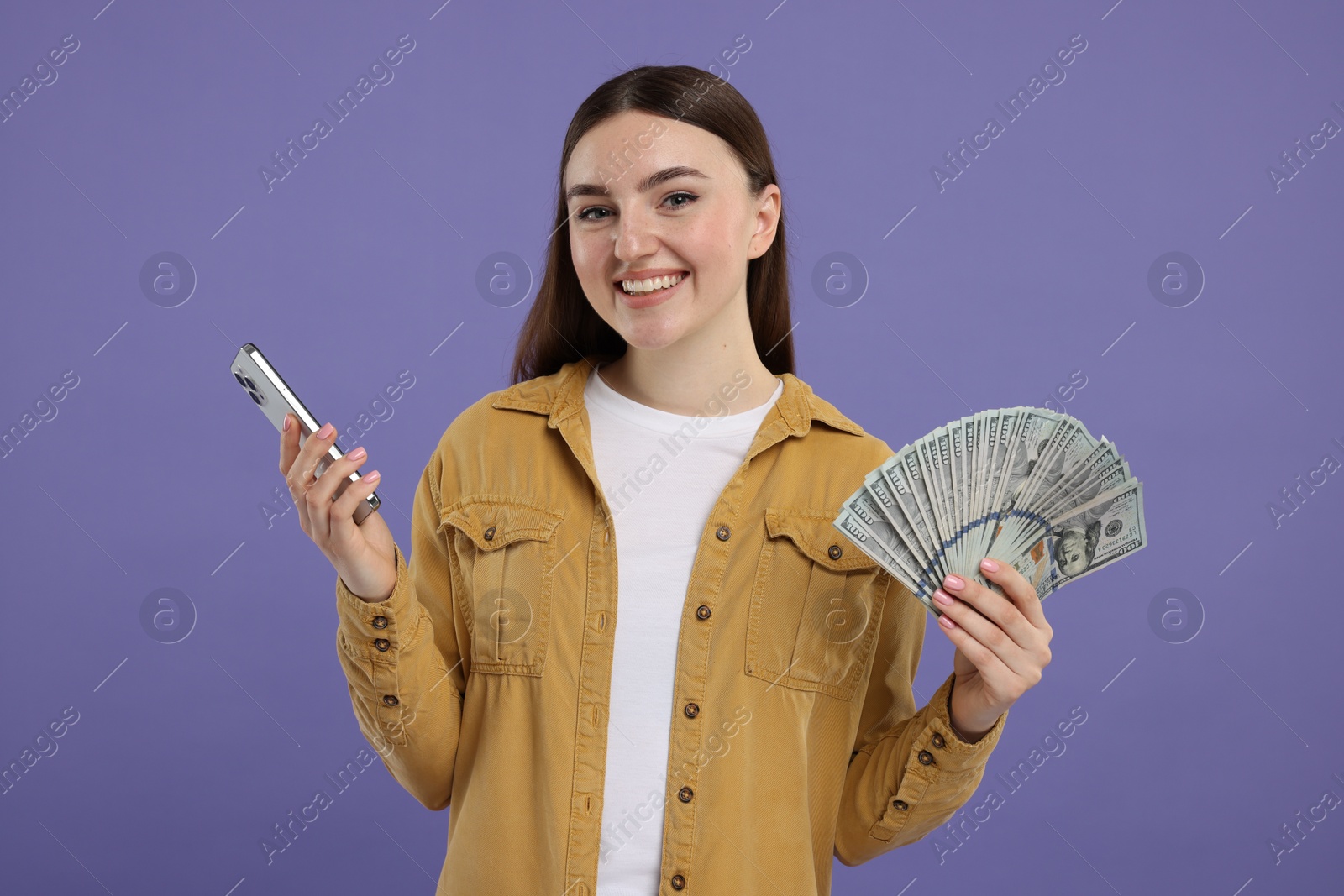 Photo of Happy woman with dollar banknotes and smartphone on purple background