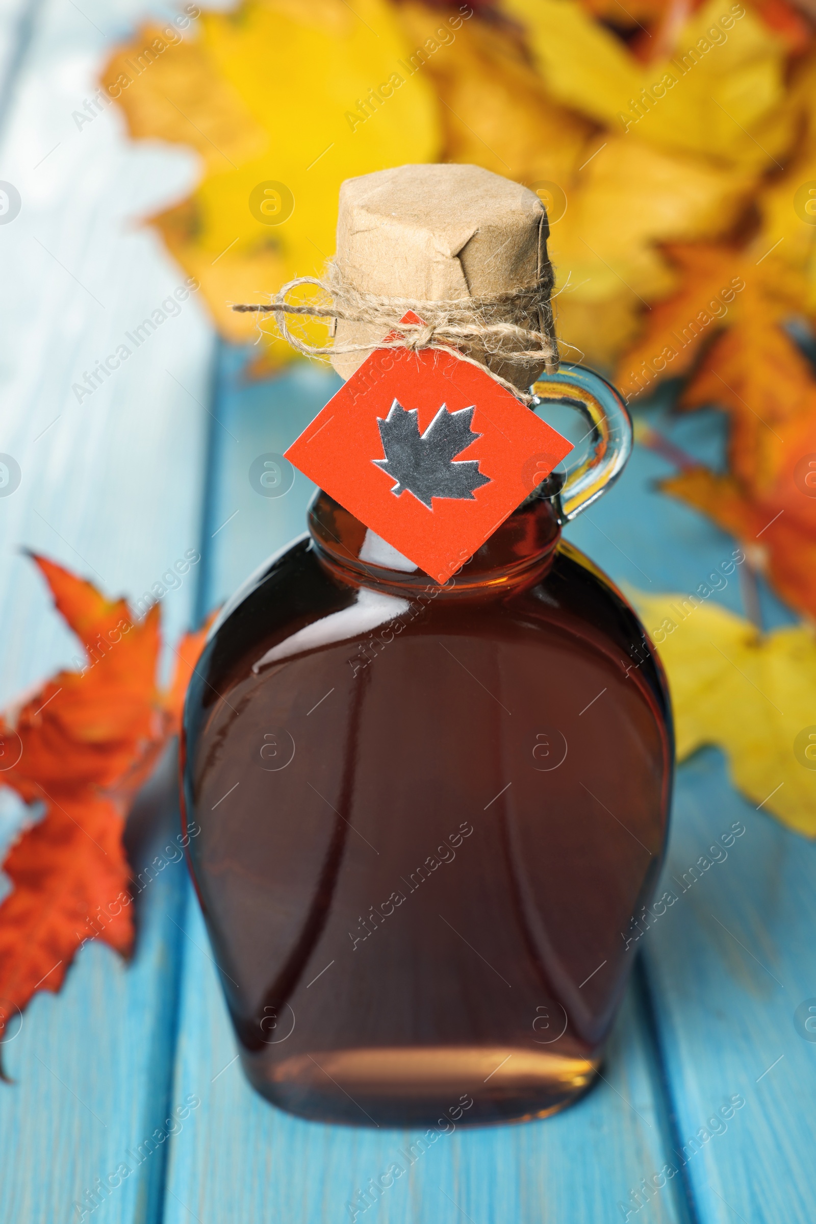 Photo of Glass bottle of tasty maple syrup and dry leaves on light blue wooden table, closeup