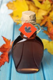 Photo of Glass bottle of tasty maple syrup and dry leaves on light blue wooden table, closeup