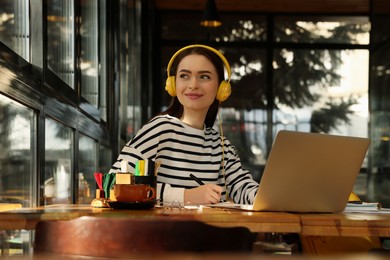 Young female student with laptop and headphones studying at table in cafe