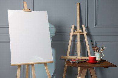Photo of Easel with blank canvas, cup of drink and different art supplies on wooden table near grey wall