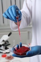 Scientist dripping red liquid into Petri dish with raw cultured meat in laboratory, closeup
