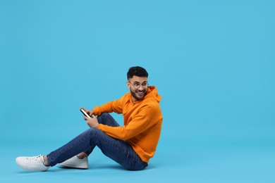 Happy young man using smartphone on light blue background, space for text