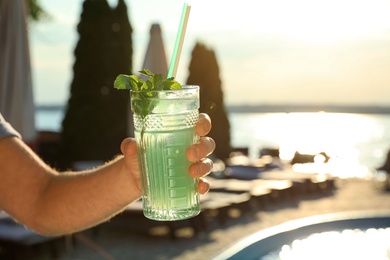 Photo of Man with glass of fresh summer cocktail near swimming pool outdoors at sunset, closeup. Space for text
