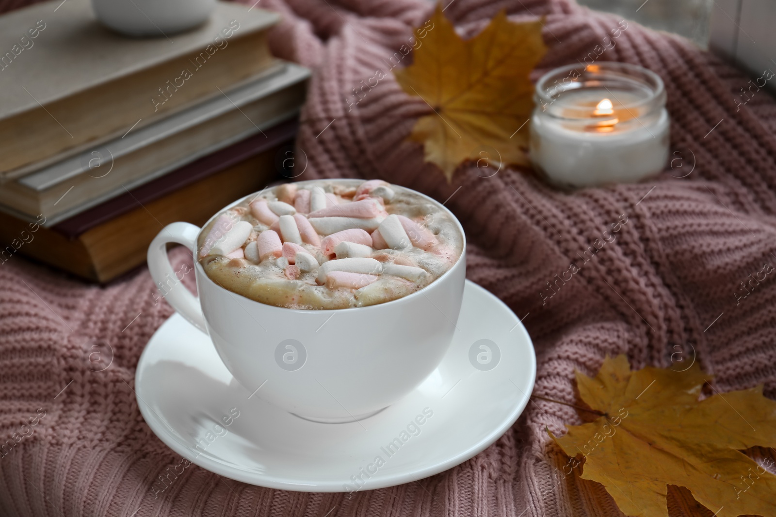 Photo of Cup of tasty hot drink with marshmallows, books, leaves and burning candle on blanket. Autumn coziness