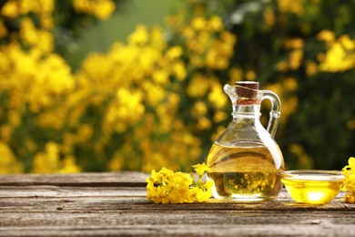 Photo of Rapeseed oil in glass jug, bowl and beautiful yellow flowers on wooden table outdoors, space for text