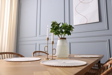 Vase with green branches and burning candles on wooden table in stylish dining room