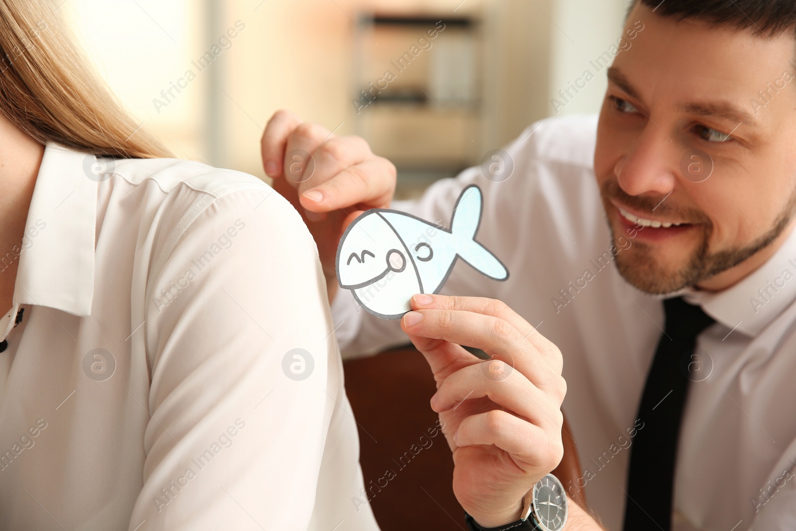 Photo of Man sticking paper fish to colleague's back in office. Funny joke