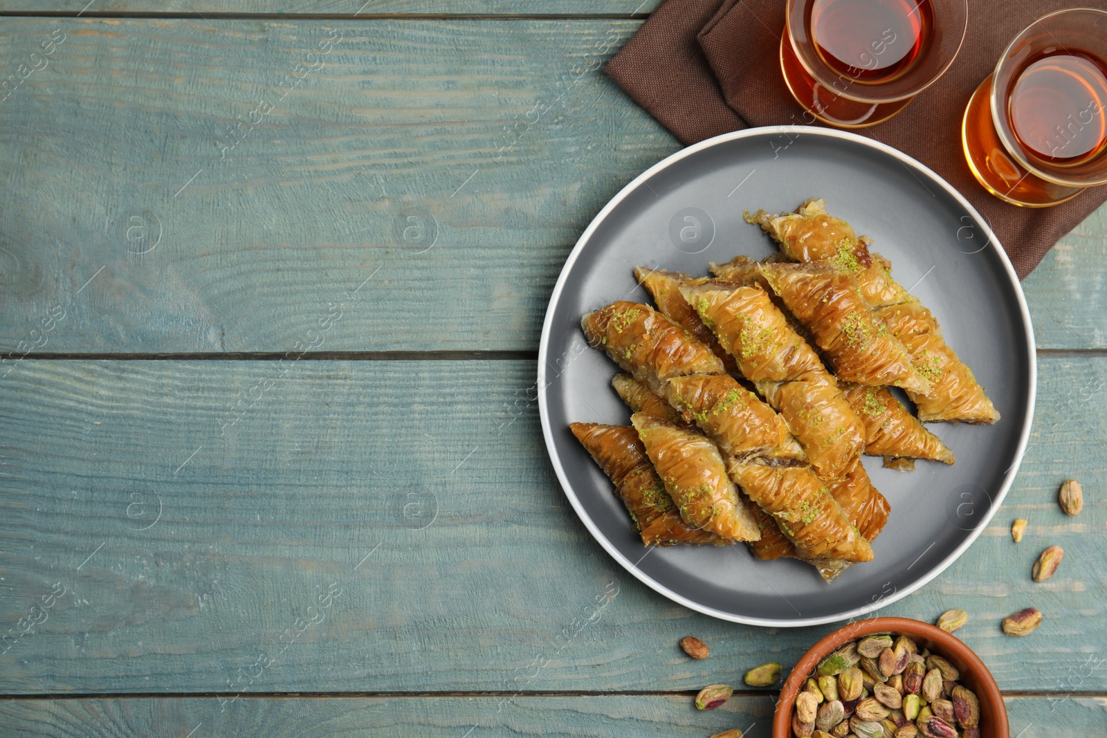 Photo of Delicious baklava with pistachios, scattered nuts and hot tea on light blue wooden table, flat lay. Space for text