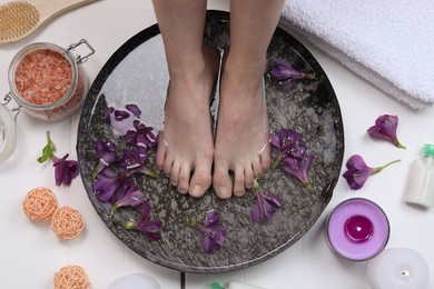 Photo of Woman soaking her feet in bowl with water and flowers on white wooden floor, top view. Spa treatment