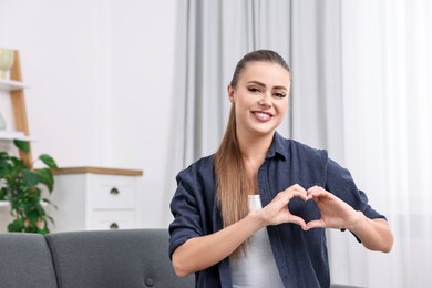 Happy woman showing heart gesture with hands at home, space for text