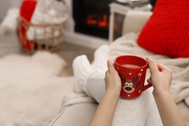 Woman with cup of hot drink resting on sofa near fireplace in living room, closeup