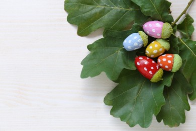 Photo of Colorful painted acorns with polka dot pattern and green oak leaves on white wooden table, flat lay. Space for text