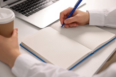 Young woman with cup of coffee writing in notebook at white table, closeup