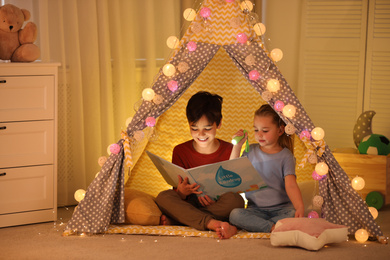 Little children with flashlight reading book in play tent at home