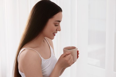 Happy woman with cup of drink near window indoors, space for text. Lazy morning
