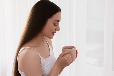 Photo of Happy woman with cup of drink near window indoors, space for text. Lazy morning