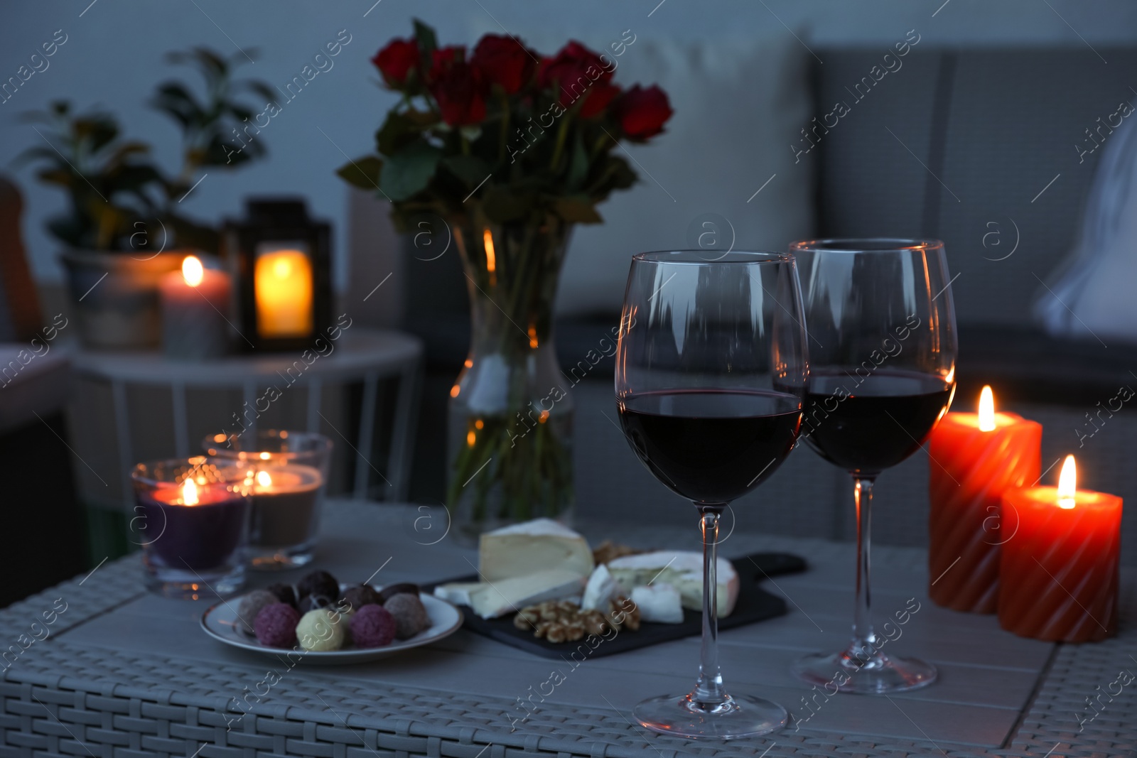 Photo of Glasses of wine, vase with roses, burning candles and snacks on rattan table at balcony in evening, closeup