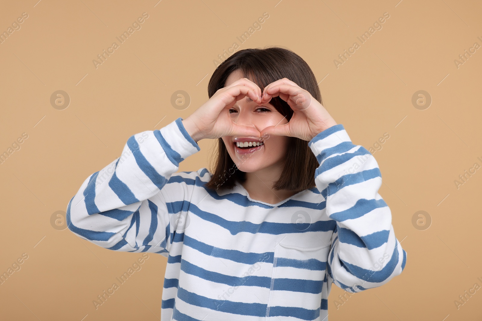 Photo of Happy woman looking through folded in shape of heart hands on beige background
